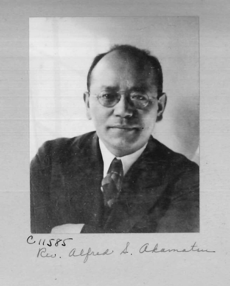 Black and white picture of a smiling Japanese man. He has short hair and glasses, and is wearing a suit.