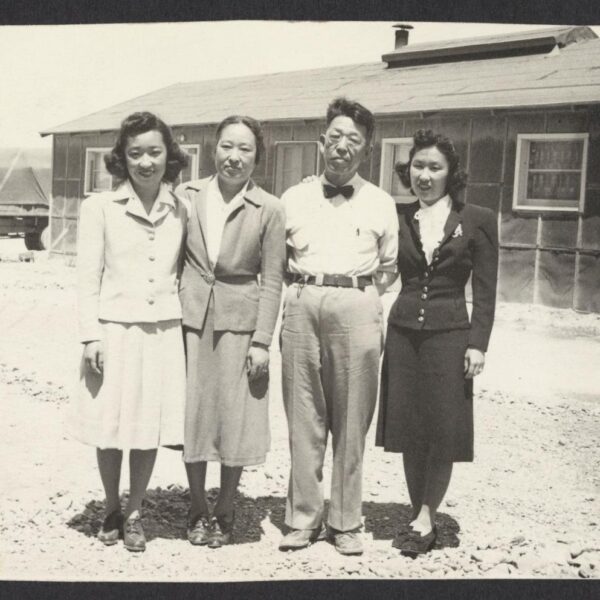Black and white photograph of a family of four Japanese people, three women and a man, in front of a bunk house at the Topaz internment camp.