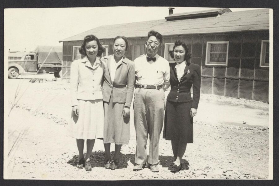 Black and white photograph of a family of four Japanese people, three women and a man, in front of a bunk house at the Topaz internment camp.
