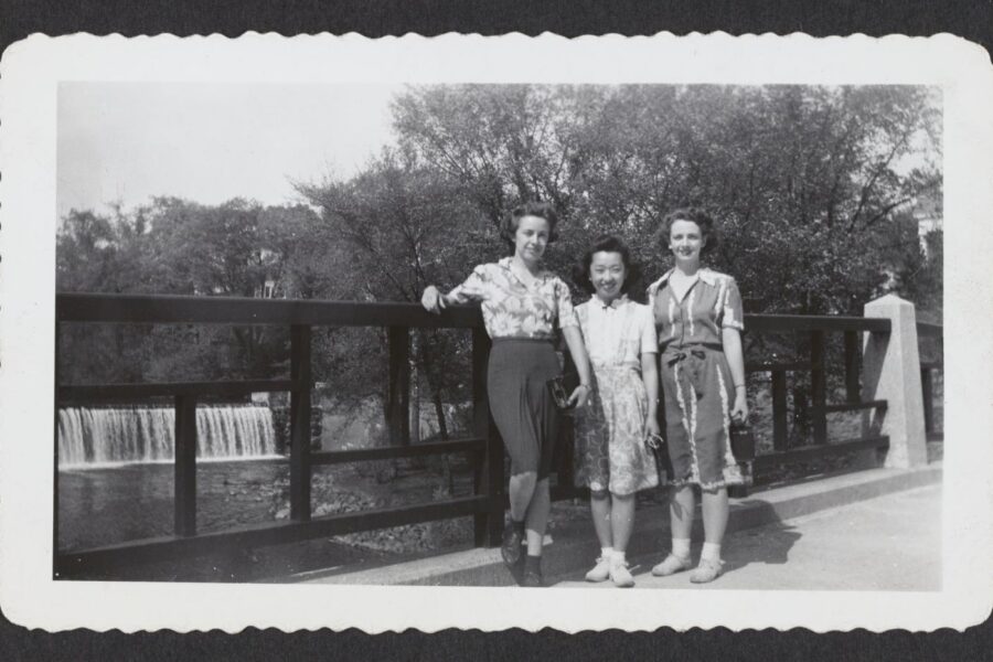 A black and white photograph of three women standing on a bridge in front of a small waterfall.