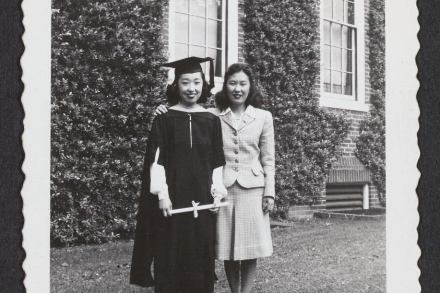 A black and white photograph of two Japanese women, left, Yoshiko Uchida in graduation garb, standing in front of a foliage covered brick wall with windows.