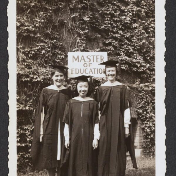 Black and white photo of three women in graduation garb standing in front of a sign that reads 