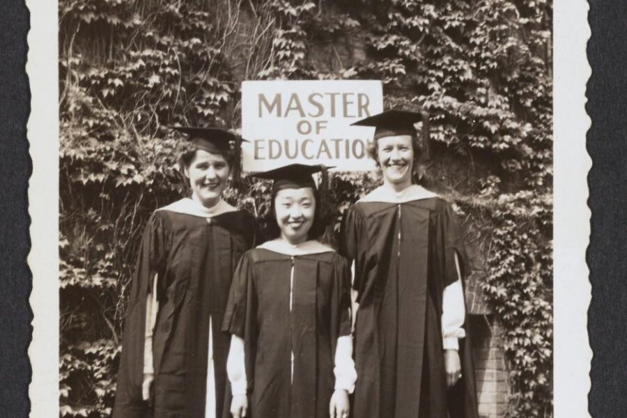Black and white photo of three women in graduation garb standing in front of a sign that reads 
