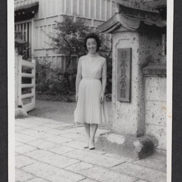 A black and white photograph of Yoshiko Uchida, a Japanese American woman, standing in front of the entrance marker for Nihon Mingei Kai.