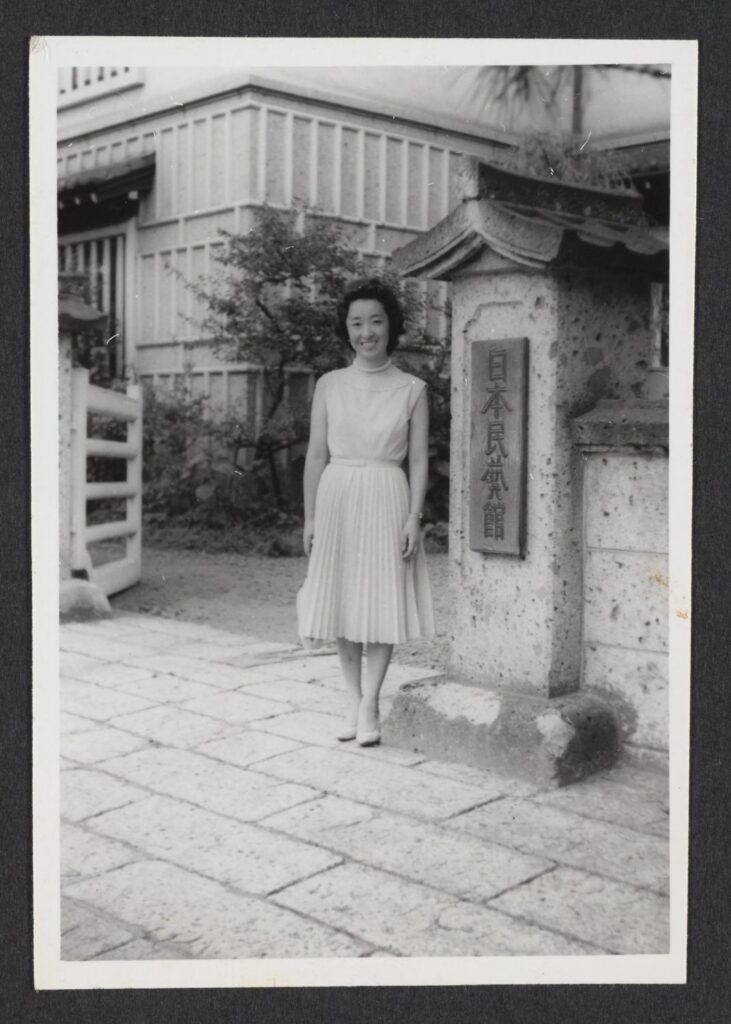 A black and white photograph of Yoshiko Uchida, a Japanese American woman, standing in front of the entrance marker for Nihon Mingei Kai.