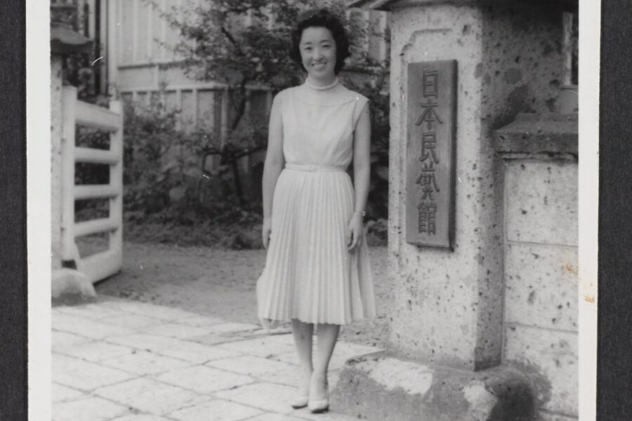 A black and white photograph of Yoshiko Uchida, a Japanese American woman, standing in front of the entrance marker for Nihon Mingei Kai.