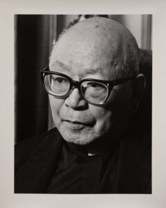 A black and white photo of an older Japanese man, bald, with thick black glasses. He is looking slightly down and to the left.
