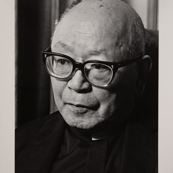 A black and white photo of an older Japanese man, bald, with thick black glasses. He is looking slightly down and to the left.