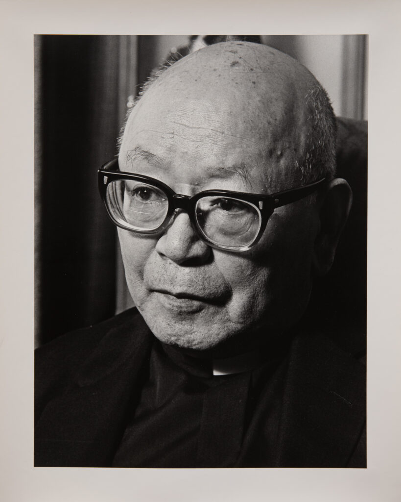 A black and white photo of an older Japanese man, bald, with thick black glasses. He is looking slightly down and to the left.