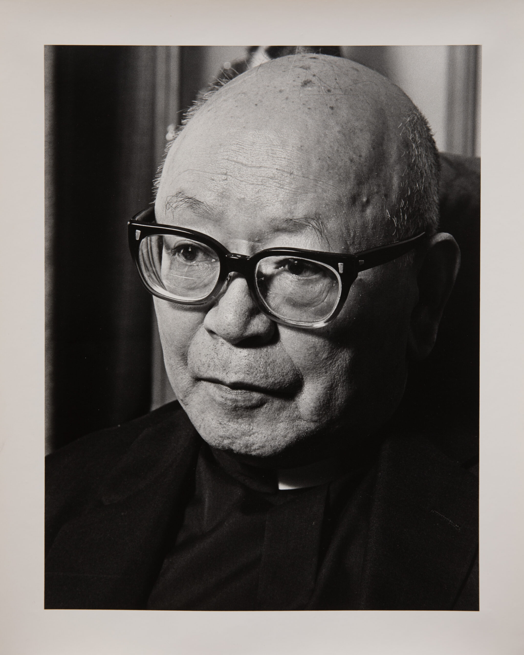 A black and white photo of an older Japanese man, bald, with thick black glasses. He is looking slightly down and to the left.