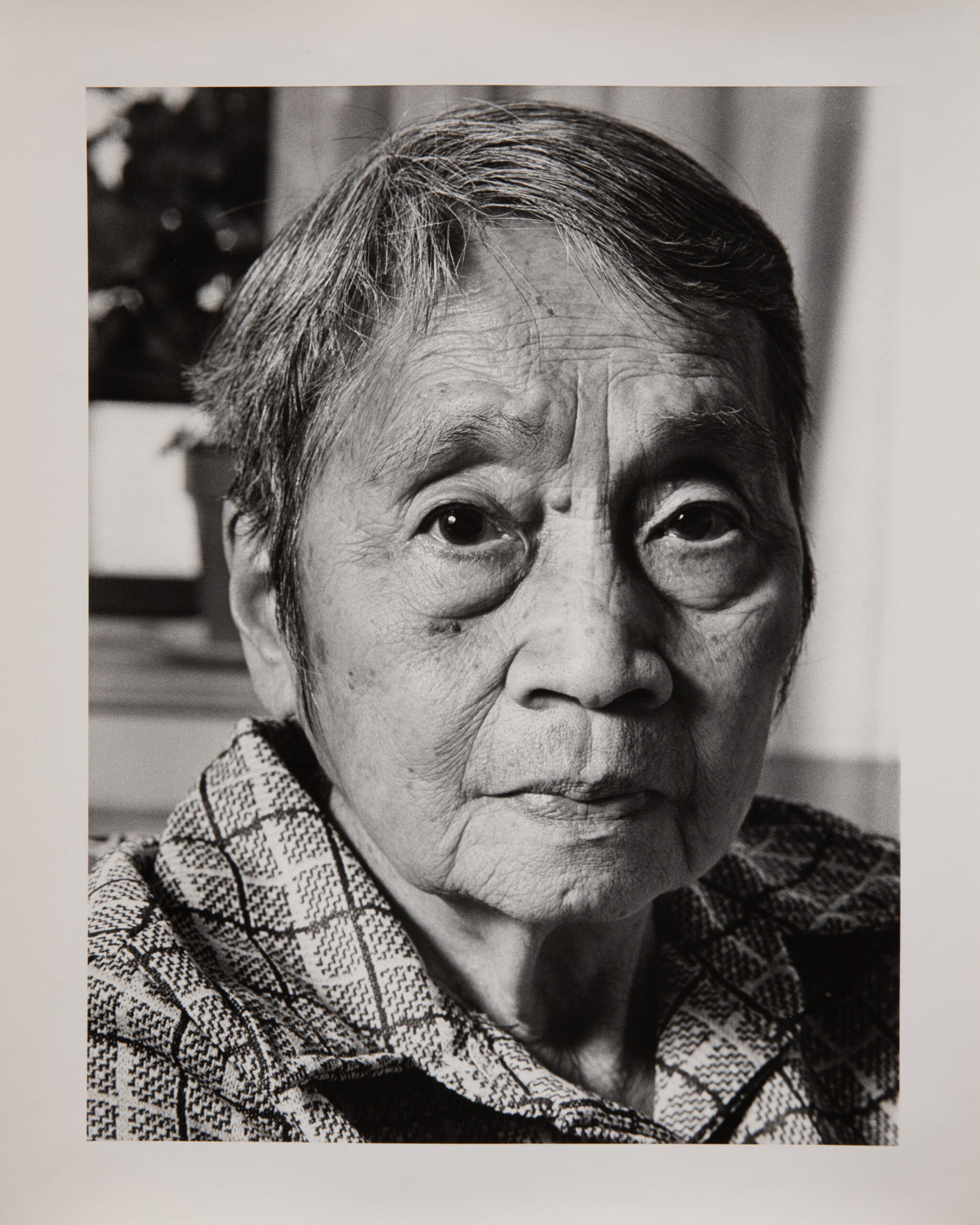 A black and white portrait photograph of an older Japanese woman. She has short hair. Her expression is serious and she is looking straight at the camera. She wears a striped button down shirt.
