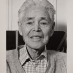 A black and white portrait photo of an older Japanese man. He is wearing a plaid collared shirt under a cardigan. He has white hair. He is looking directly at the camera and smiling faintly.