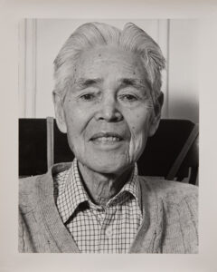 A black and white portrait photo of an older Japanese man. He is wearing a plaid collared shirt under a cardigan. He has white hair. He is looking directly at the camera and smiling faintly.