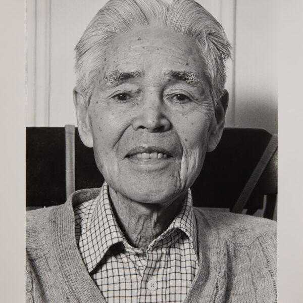 A black and white portrait photo of an older Japanese man. He is wearing a plaid collared shirt under a cardigan. He has white hair. He is looking directly at the camera and smiling faintly.