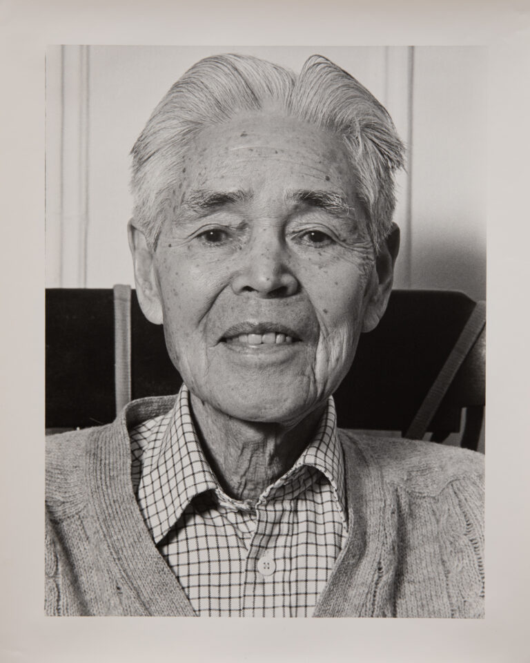 A black and white portrait photo of an older Japanese man. He is wearing a plaid collared shirt under a cardigan. He has white hair. He is looking directly at the camera and smiling faintly.
