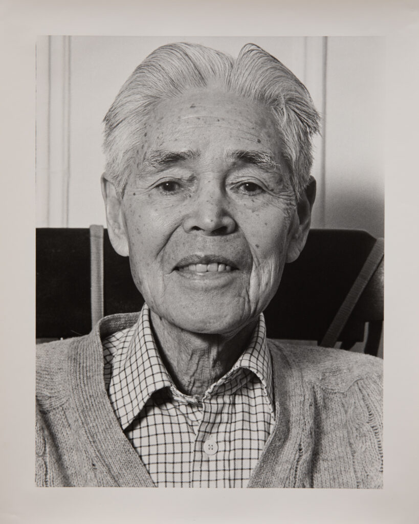 A black and white portrait photo of an older Japanese man. He is wearing a plaid collared shirt under a cardigan. He has white hair. He is looking directly at the camera and smiling faintly.