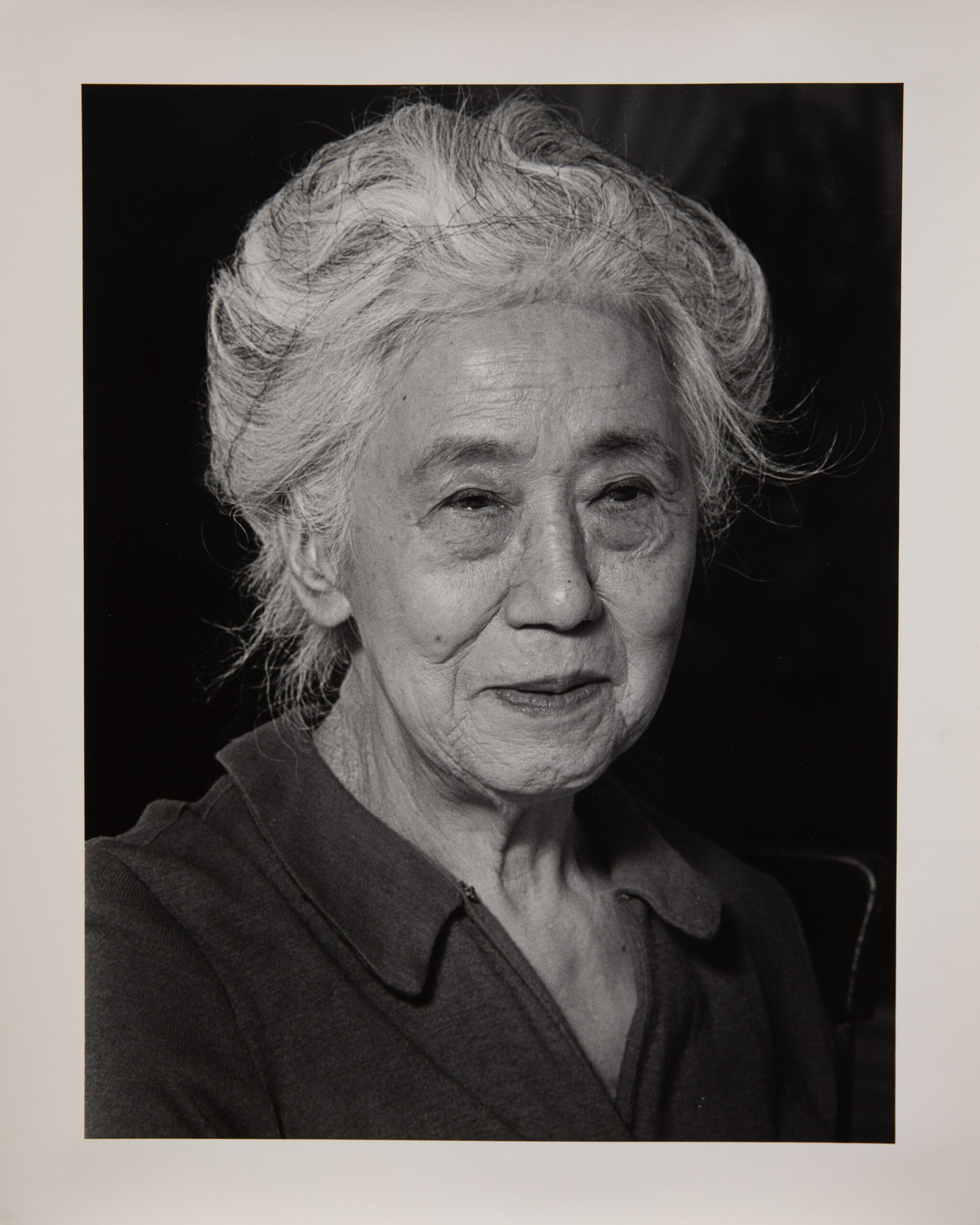 A black and white portrait photograph of an older Japanese woman. She is wearing a casual v-neck shirt with a collar. She has white hair that is held up with a hair net. She is looking to the right and not directly at the camera. She has a faint smile on her lips.