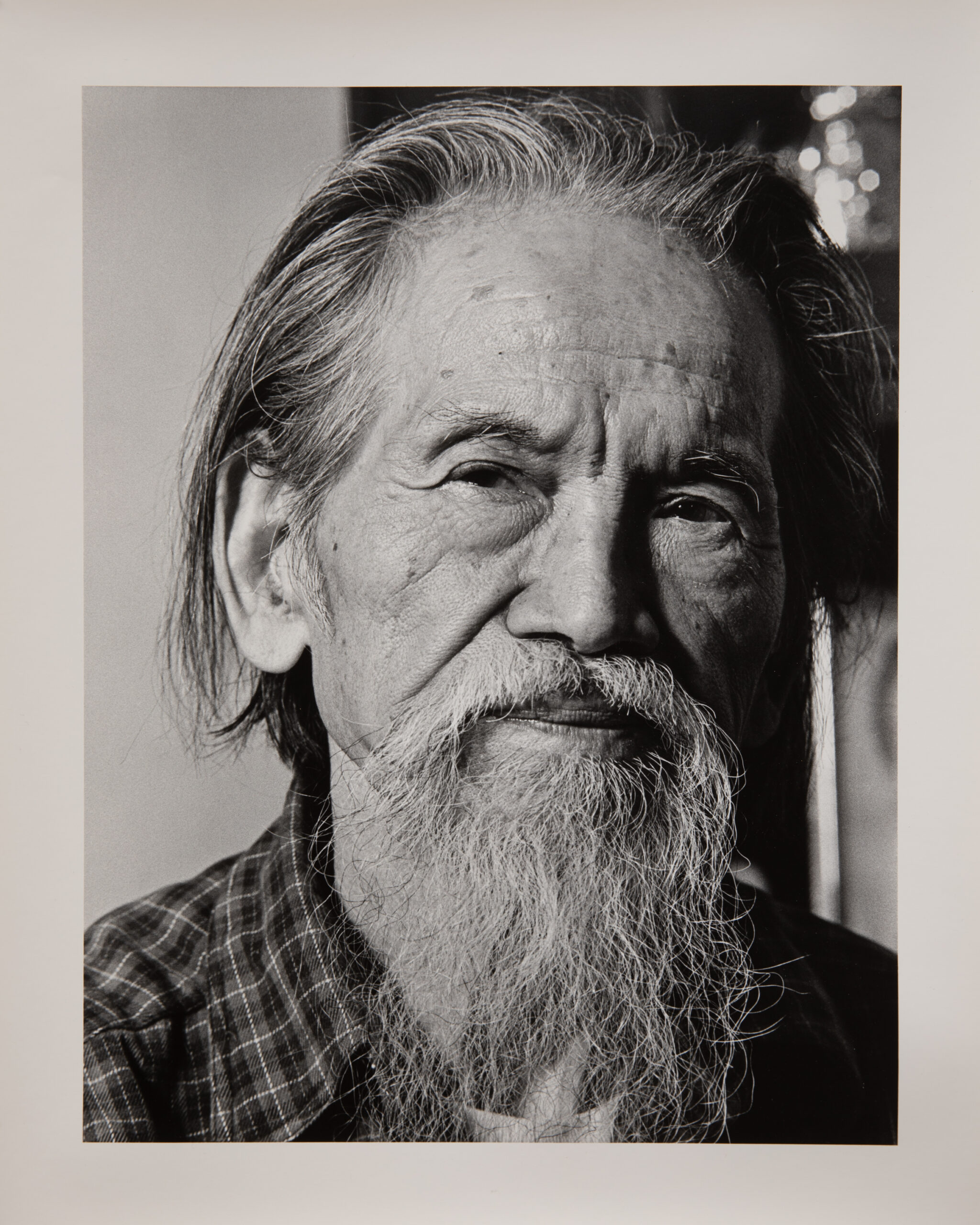 A black and white portrait photograph of an older Japanese man. He is wearing a plaid shirt over a white t-shirt. His hair is longer and greying, and he has a long grey beard. He has a slight smile is looking at the camera from a slight angle.